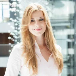 Portret uśmiechniętej kobiety z długimi blond włosami w białej koszuli na tle nowoczesnego biura z szklanymi elementami i dekoracyjnym oświetleniem.