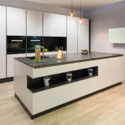 Nowoczesna kuchnia z wyspą i białymi frontami, czarne akcenty, blat z ciemnego kamienia, dekoracyjne lampy wiszące, słoik makaronu i drewniany blok na noże na blacie.