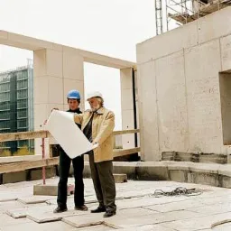 Dwóch mężczyzn w kaskach analizuje plan architektoniczny na placu budowy, widoczne surowe betonowe elementy konstrukcyjne i rusztowania w tle.