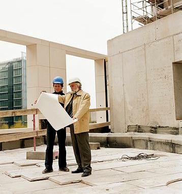 Dwóch mężczyzn w kaskach analizuje plan architektoniczny na placu budowy, widoczne surowe betonowe elementy konstrukcyjne i rusztowania w tle.