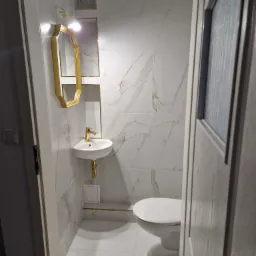 Mała, biała łazienka z marmurowymi płytkami, złotą armaturą i lustrem w ozdobnej, złotej ramie, widoczna zza uchylonych drzwi.