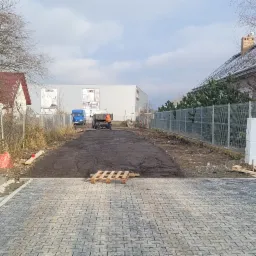 Układanie kostki brukowej na nowym podjeździe, widoczny fragment utwardzonej drogi gruntowej oraz ciężarówka z pracownikiem w pomarańczowej kamizelce w tle.