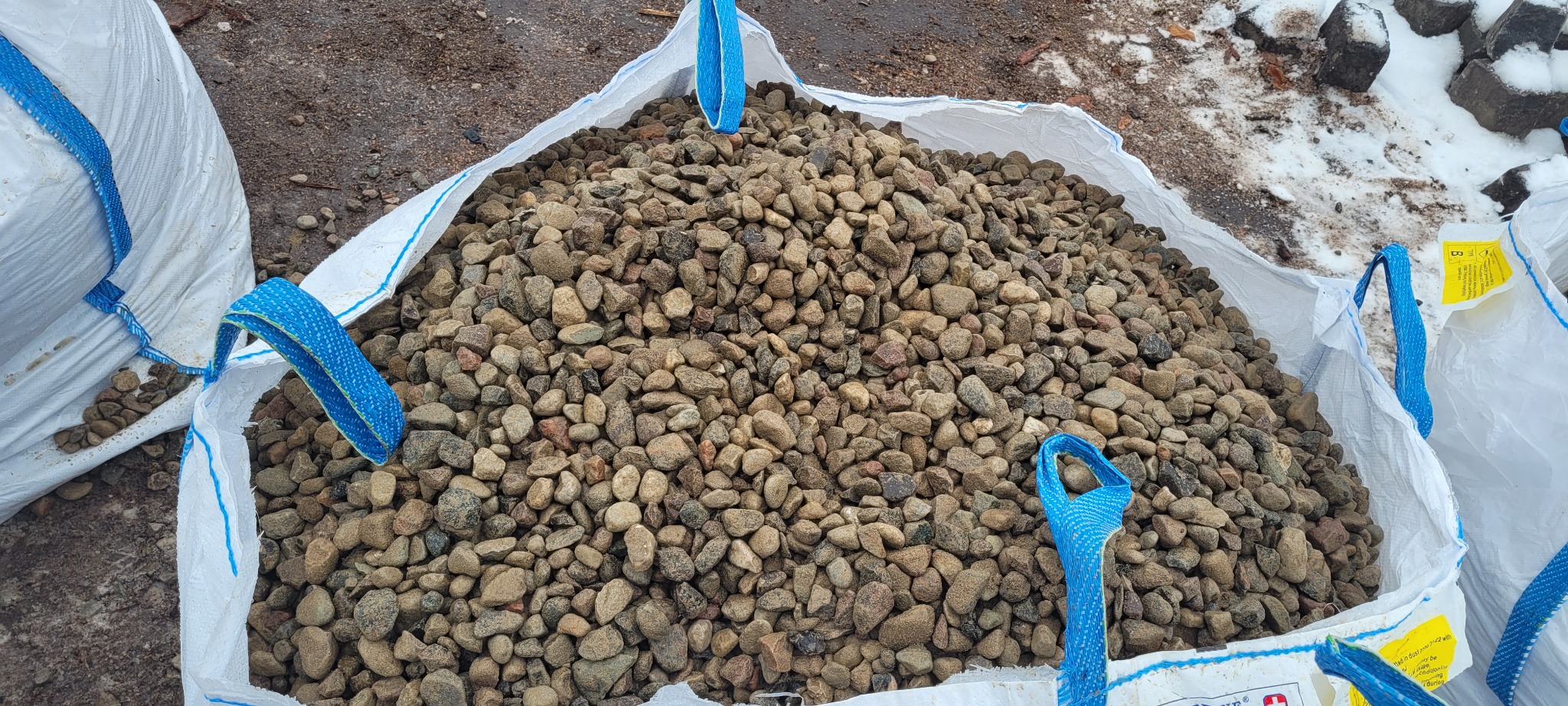 Worek typu big bag wypełniony brązowo-szarym kruszywem, widoczne niebieskie uchwyty worka, w tle fragmenty śniegu i ziemi.