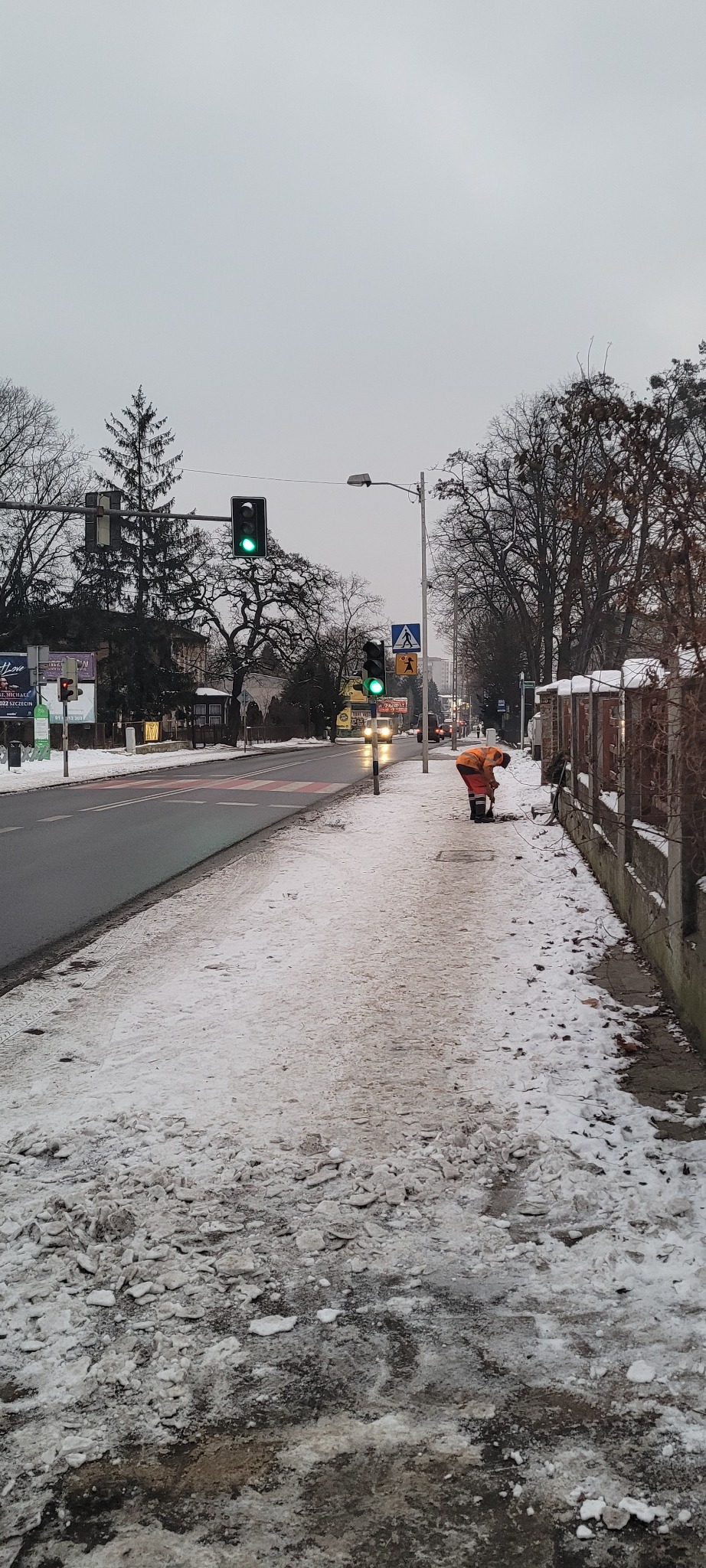Pracownik w pomarańczowym kombinezonie odśnieża chodnik przy ulicy w pochmurny dzień, widoczne resztki śniegu i lodu na chodniku i jezdni.