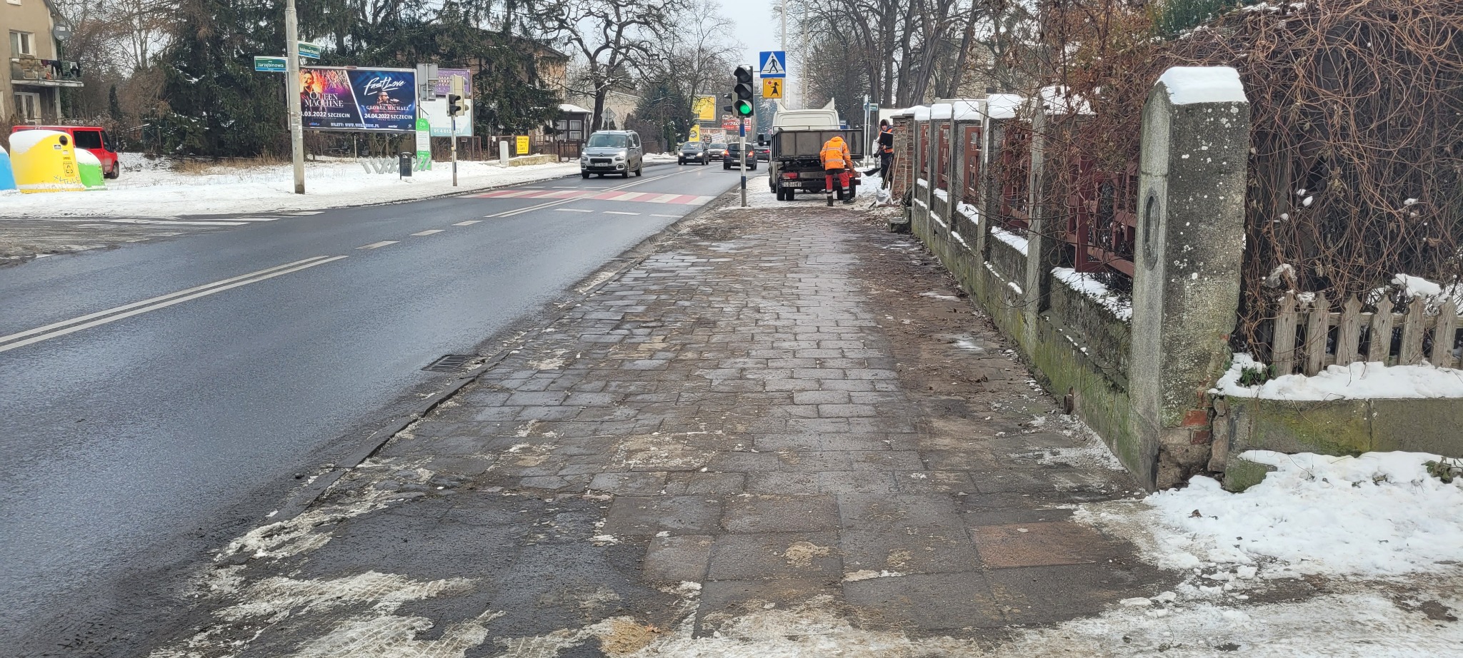 Dwaj pracownicy w pomarańczowych kombinezonach odśnieżają chodnik miejski przy ulicy w zimowy dzień. Na chodniku leży śnieg i błoto pośniegowe, a w tle widać samochody i budynki.