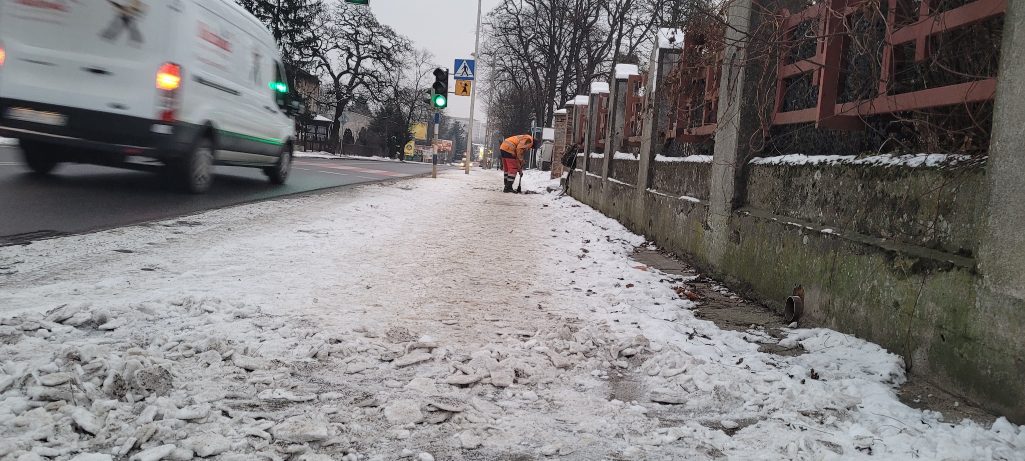 Pracownik w pomarańczowej kamizelce odblaskowej odśnieża chodnik łopatą, obok przejeżdża biały samochód dostawczy, w tle sygnalizacja świetlna i budynki.