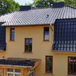 Dwupiętrowy dom z nowym, ciemnoszarym dachem z dachówki ceramicznej, widoczne rynny i obróbki blacharskie, fragment dachu werandy w trakcie prac wykończeniowych.