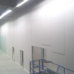 Firma Ogólnobudowlana Marcin Lichosik - Montaż ściany działowej z płyt karton-gips w hali magazynowej, widoczne łączenia płyt i metalowa konstrukcja otworów drzwiowych, oświetlenie sufitowe.