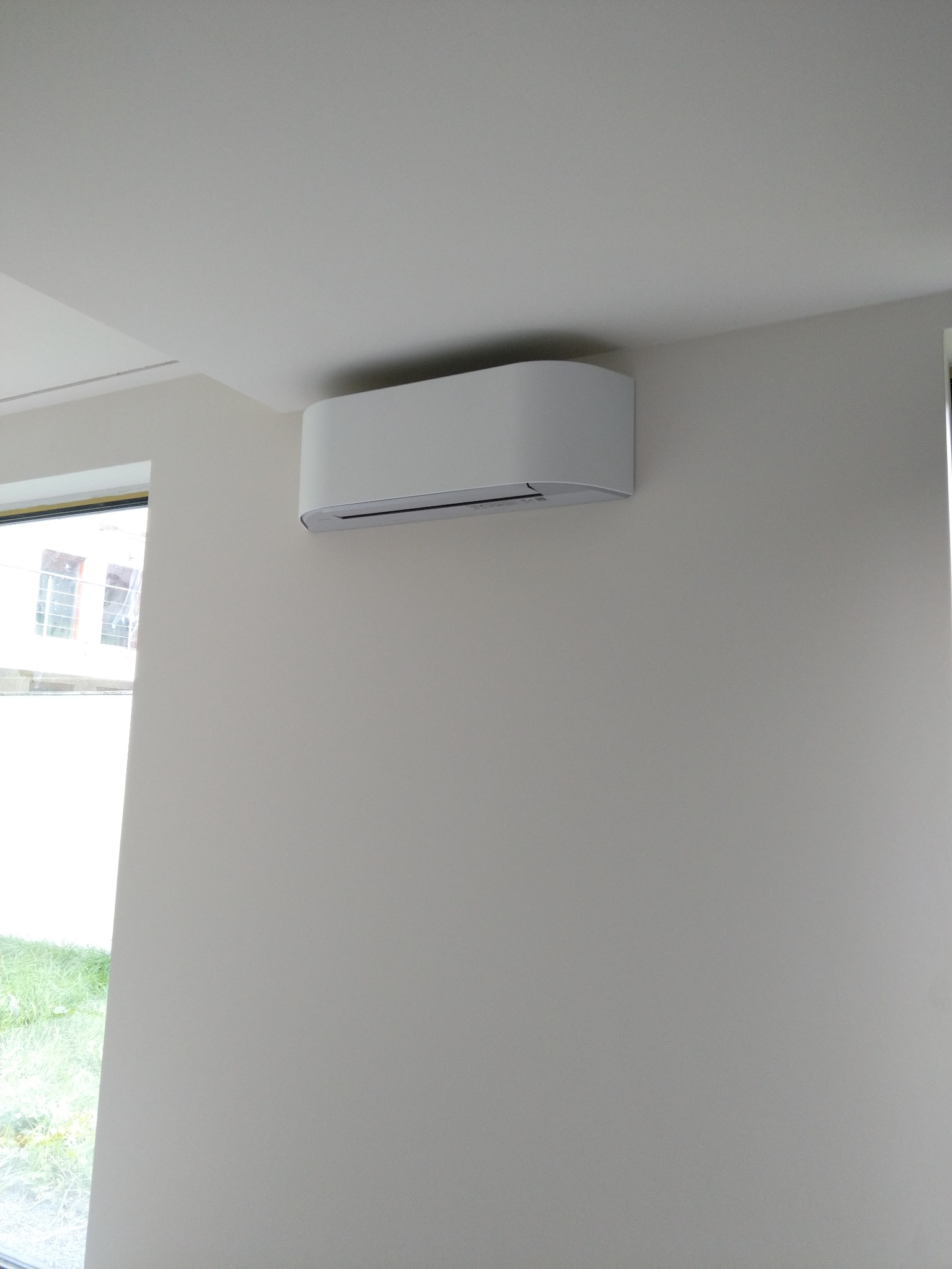 Biały klimatyzator ścienny o zaokrąglonych kształtach w jasnym wnętrzu. Widok z dołu, okno z widokiem na zieleń w tle.