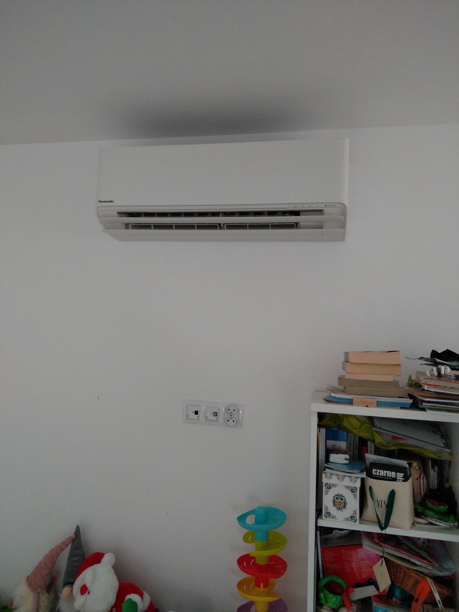Biały klimatyzator Panasonic zamontowany na białej ścianie w pokoju dziecięcym, widoczne gniazdka i regał z zabawkami.