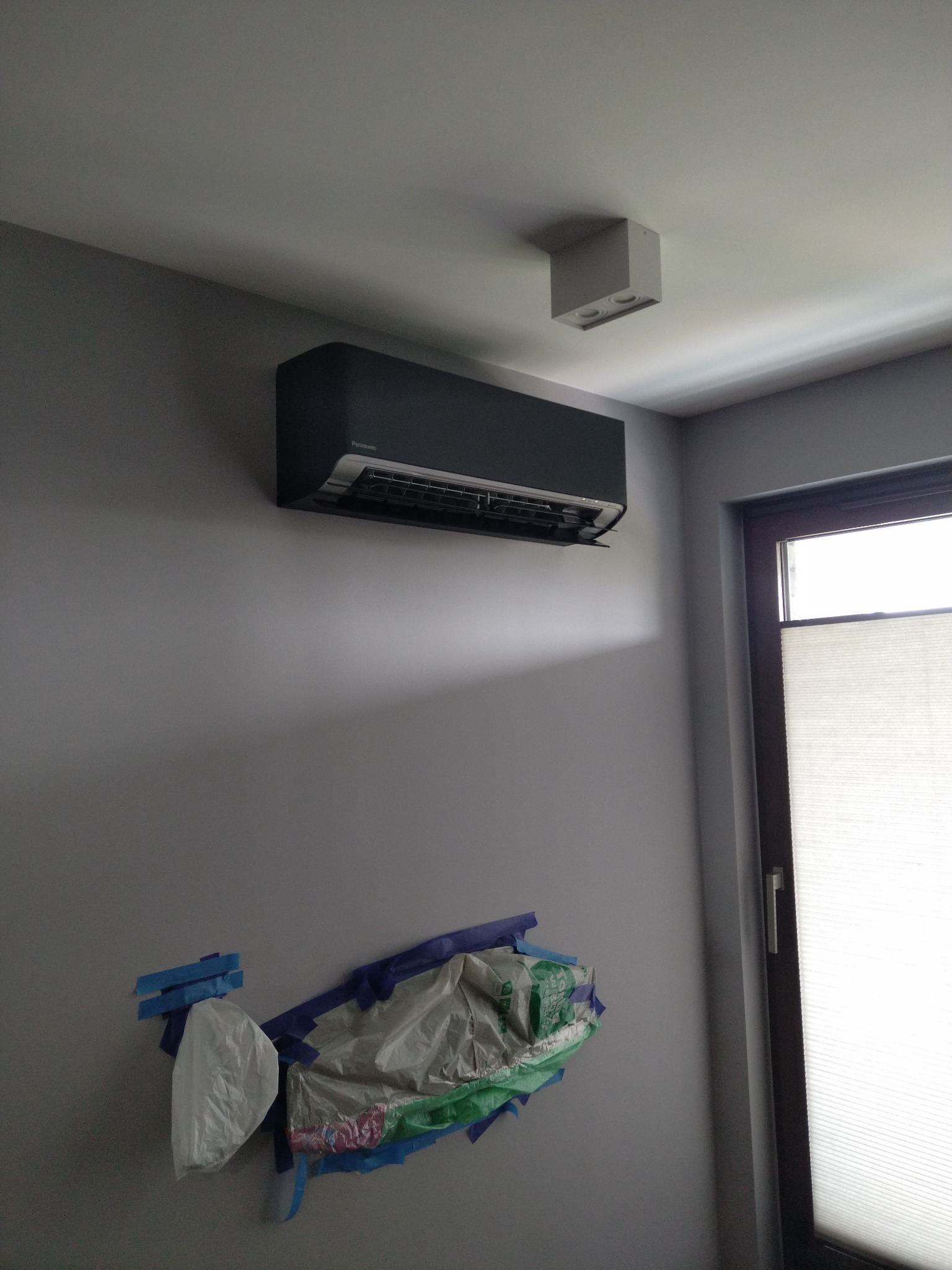 Zamontowany klimatyzator Panasonic w szarej tonacji w Rącznej, zabezpieczony folią malarską. Widoczne oświetlenie sufitowe i okno z żaluzjami.