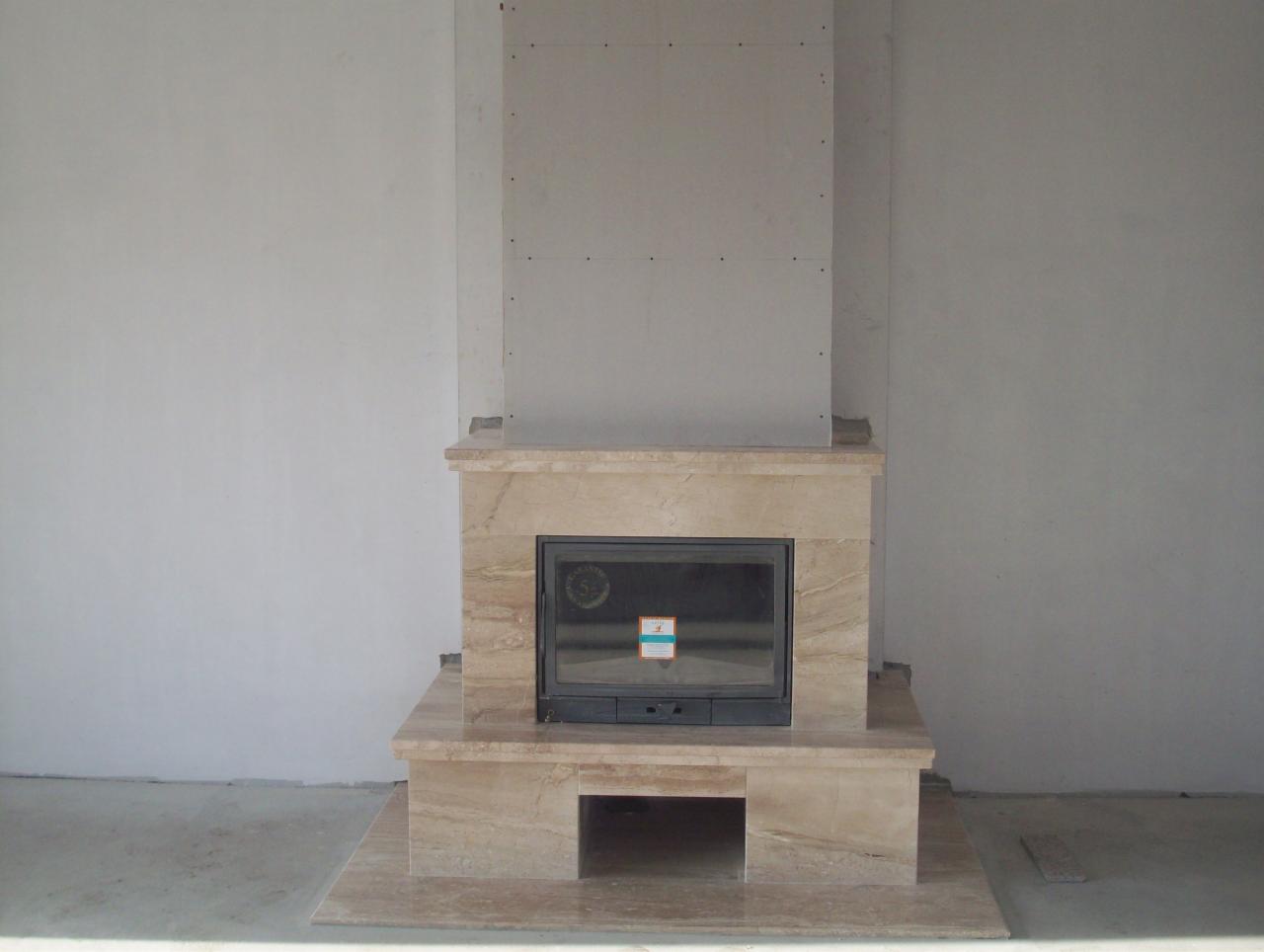 Kominek z obudową z trawertynu w trakcie montażu, widoczne płyty gipsowe na ścianie, surowy stan wykończenia wnętrza.