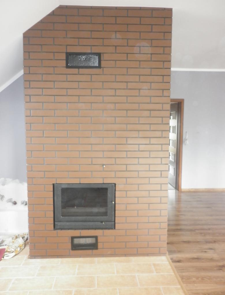 Murowany kominek z cegły klinkierowej z czarnym wkładem kominkowym i kratkami wentylacyjnymi, widoczny fragment podłogi z płytek ceramicznych i parkietu.