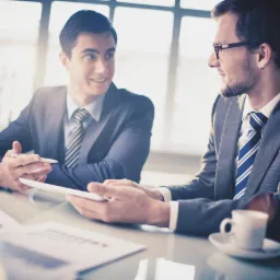 Dwóch mężczyzn w garniturach omawia dane na tablecie przy biurku z dokumentami i filiżanką kawy, w tle jasne okno.