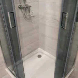 Narożna kabina prysznicowa z przesuwnymi drzwiami, chromowana armatura i odpływ, płytki imitujące marmur na ścianach.