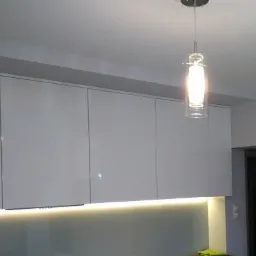 Nowoczesna kuchnia z białymi, lakierowanymi szafkami wiszącymi, oświetleniem LED podszafkowym i szklanym kloszem lampy wiszącej z żarówką.