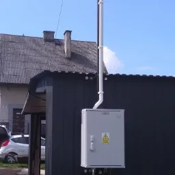 Zewnętrzna skrzynka elektryczna z symbolem ostrzegawczym, zamontowana na ścianie budynku, z pionowym przewodem i uziemieniem, oraz przewodem odgromowym na słupie