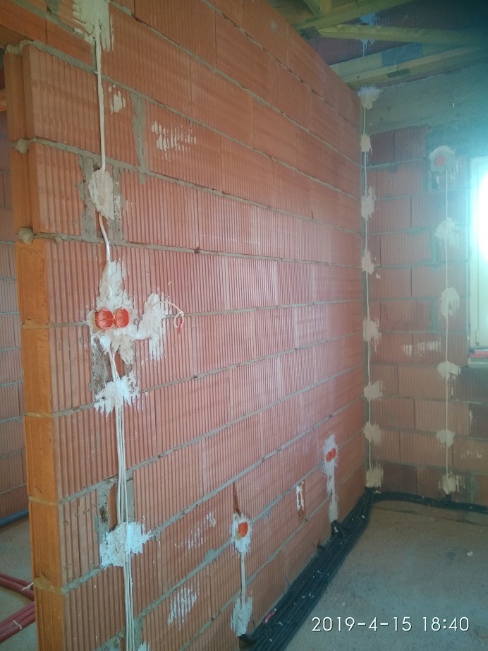 Murowana ściana z pustaków z rozprowadzoną instalacją elektryczną, widoczne kable w białych osłonach przymocowane gipsem oraz puszki elektryczne w kolorze pomarańczowym.