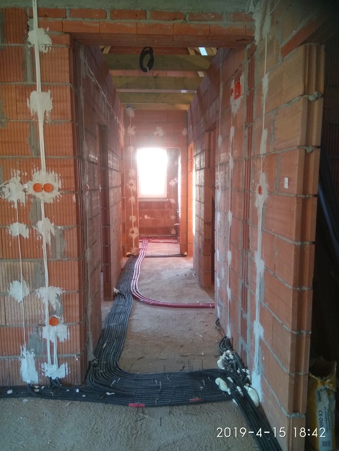 Korytarz w trakcie budowy z widocznymi przewodami elektrycznymi ułożonymi wzdłuż ścian, puszki instalacyjne zamontowane w cegłach, ślady pianki montażowej.