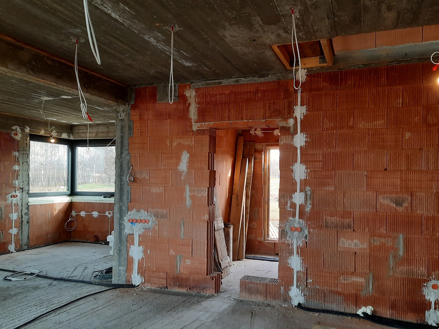Wnętrze w stanie surowym z widocznymi przewodami elektrycznymi wyprowadzonymi ze ściany i sufitu, puszki instalacyjne zamontowane w ścianach z cegły ceramicznej, przewody częściowo zamocowane gipsem.