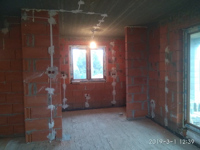 Wewnętrzne okablowanie elektryczne w surowym stanie, kable prowadzone po ceglanych ścianach, widoczne puszki instalacyjne i przewody mocowane gipsem.