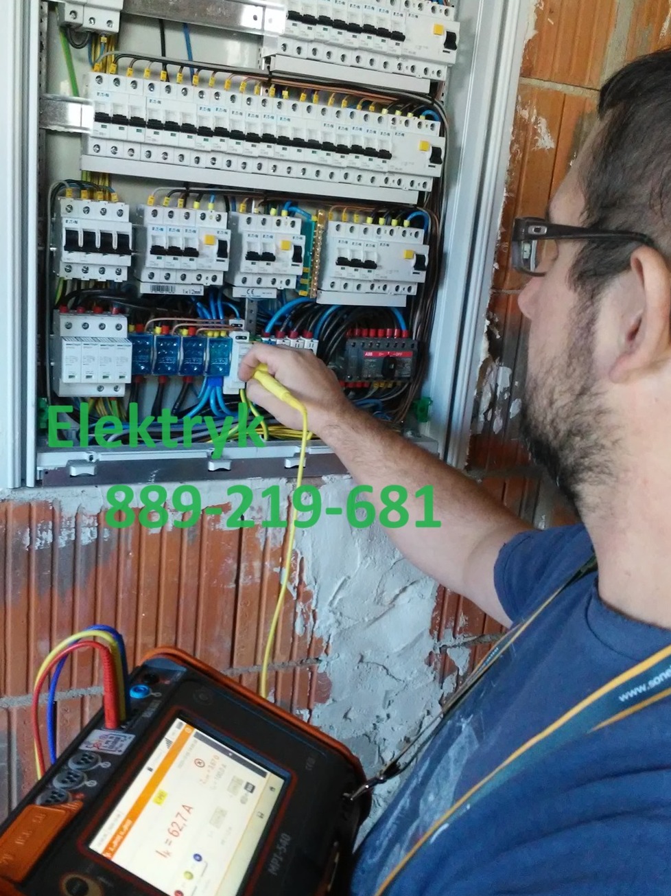 Elektryk w okularach, mierzący napięcie w skrzynce rozdzielczej w trakcie montażu, widoczny miernik z wartością 62.7A, ściana z czerwonej cegły.