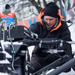 Mężczyzna w czarnej kurtce roboczej z pomarańczowymi akcentami i czarnej czapce, pochylony nad skomplikowanym systemem hydraulicznym maszyny w zimowym otoczeniu.