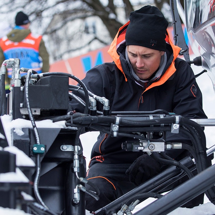 Mężczyzna w czarnej kurtce roboczej z pomarańczowymi akcentami i czarnej czapce, pochylony nad skomplikowanym systemem hydraulicznym maszyny w zimowym otoczeniu.