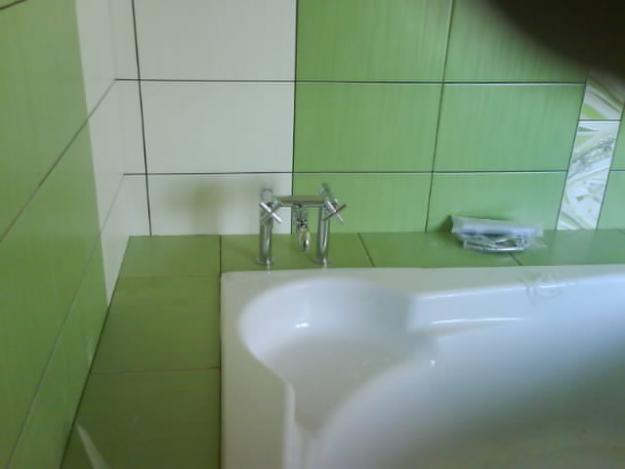 Biała wanna z chromowaną baterią na tle ściany wyłożonej zielono-białymi płytkami. Na wannie leży zapakowana mydelniczka.