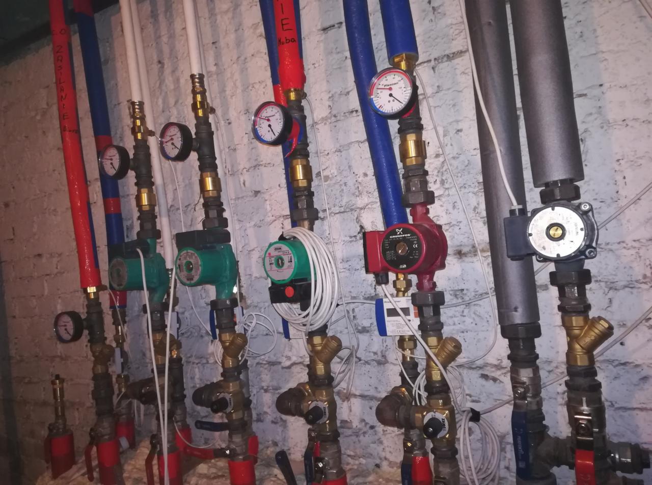 Różnorodność pomp obiegowych (czerwona, zielone) z manometrami, zamontowanych na instalacji grzewczej z rurami w kolorach czerwonym i niebieskim, na tle ściany z białej cegły.