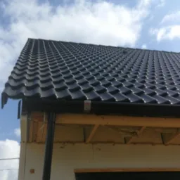 Nowy, czarny dach z blachodach&oacute;wki modułowej, widoczne rynny, fragment ocieplonej ściany budynku z zamontowanym oknem i fragment niebieskiego nieba z chmurami w tle.