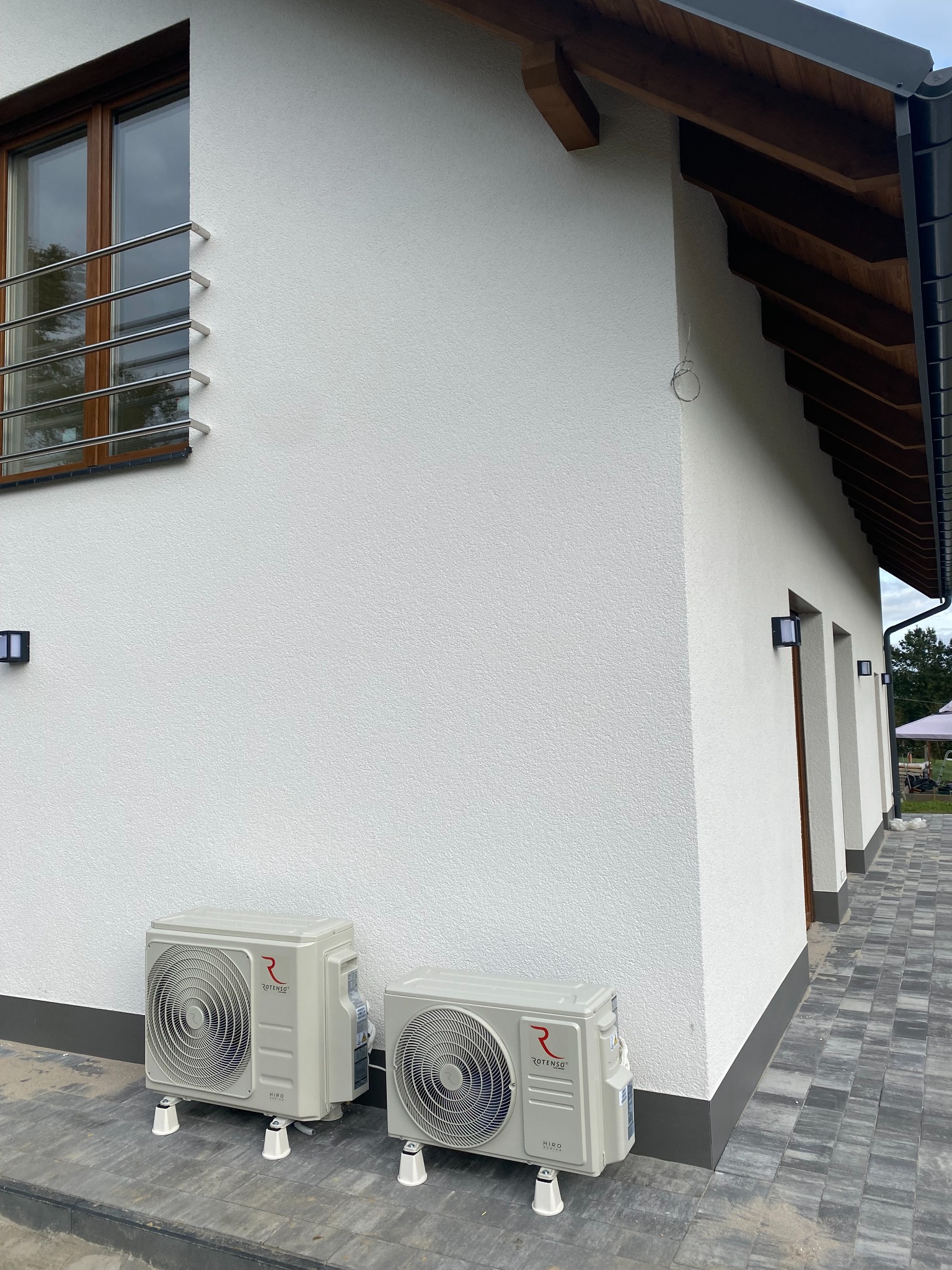 Dwa beżowe klimatyzatory Rotenso Hiro na białej elewacji domu z szarą kostką brukową, widok z boku na zainstalowane jednostki zewnętrzne.