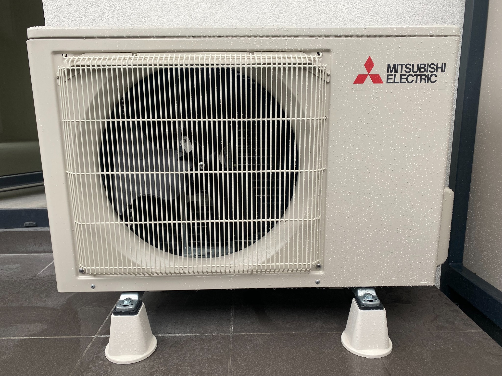 Zewnętrzna jednostka klimatyzacji Mitsubishi Electric na betonowej posadzce, pokryta kroplami wody, zamontowana na podwyższeniach.