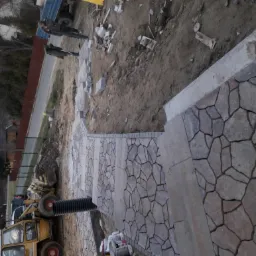 Fragment ścieżki z nieregularnej kostki granitowej w odcieniach szarości i beżu, z widocznymi elementami przygotowania terenu i sprzętem budowlanym w tle.