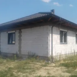 Dom w trakcie budowy z zamontowanymi oknami PCV z czarnymi ramami, widoczne białe bloczki konstrukcyjne i ciemna dachówka na dachu.