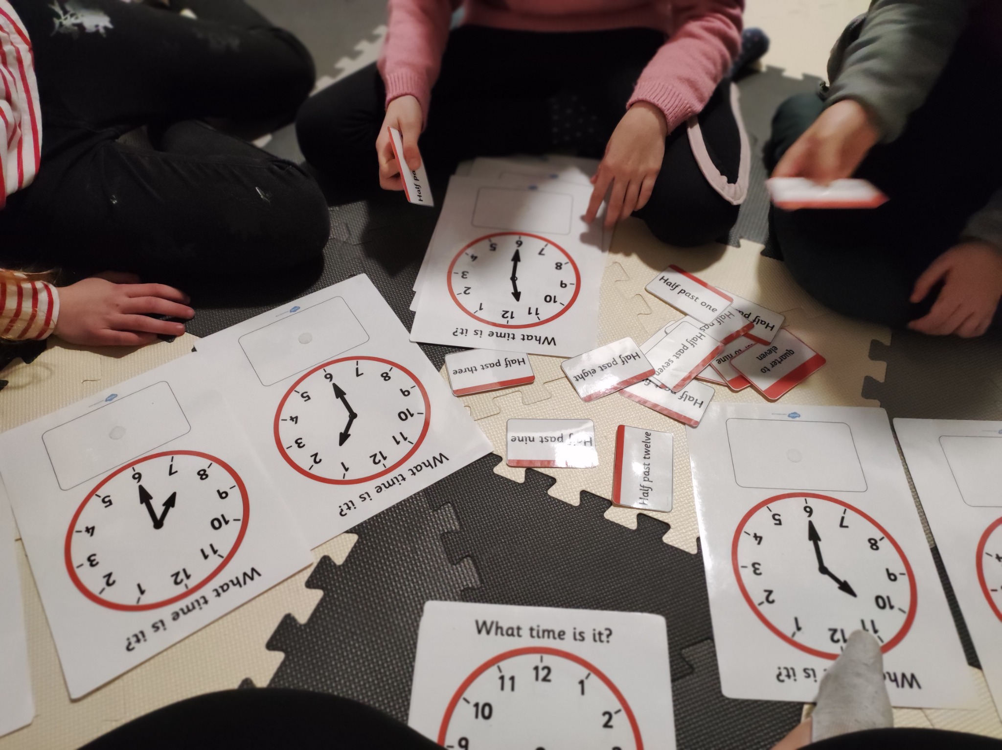 Dzieci siedzą na podłodze z puzzlami i kartami edukacyjnymi z zegarami, ucząc się odczytywania godzin.