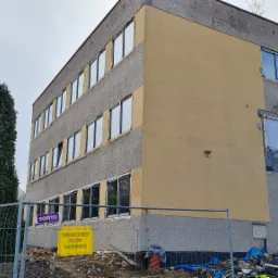 Budynek w Libiążu w trakcie wymiany okien; widoczne nowe okna oraz zabezpieczone otwory okienne, teren budowy ogrodzony.