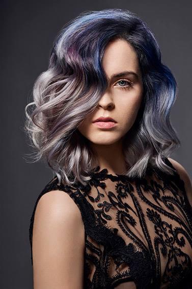 Portret kobiety z falowanymi włosami w odcieniach szarości, fioletu i granatu, z delikatnym makijażem i czarnej koronkowej sukience na ciemnoszarym tle.