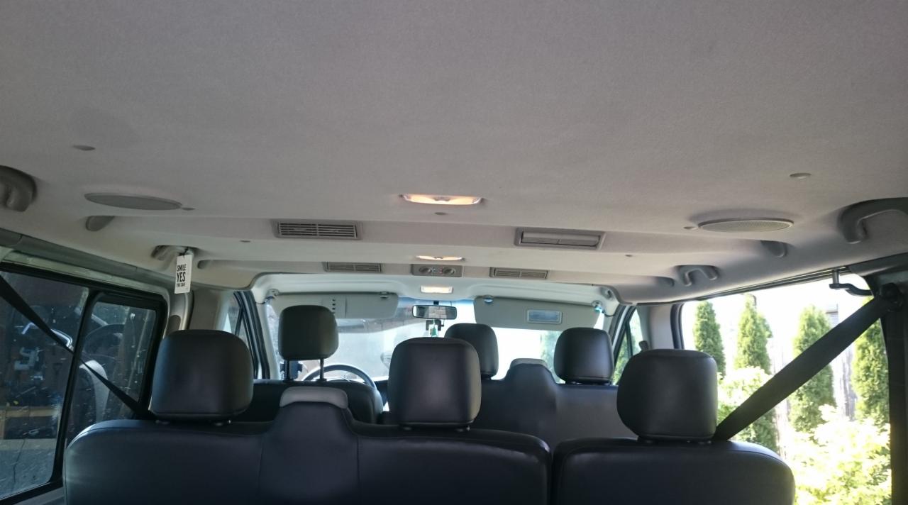 Wnętrze busa z trzema rzędami czarnych foteli, widok z tyłu kabiny, jasna podsufitka z nawiewami i oświetleniem, widoczne pasy bezpieczeństwa i zawieszka z napisem.