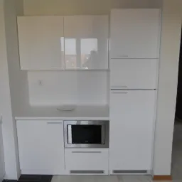 Biała zabudowa kuchenna z błyszczącymi frontami, wbudowana mikrofalówka, minimalistyczny design, talerz na blacie, widok na korytarz w tle.