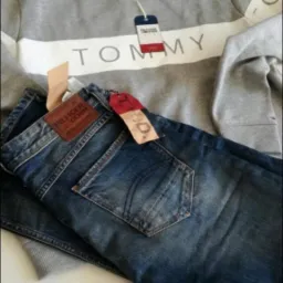 Ułożone na białym tle: szara bluza z białym pasem i napisem TOMMY oraz ciemne jeansy z brązową naszywką Hilfiger Denim i czerwoną metką.