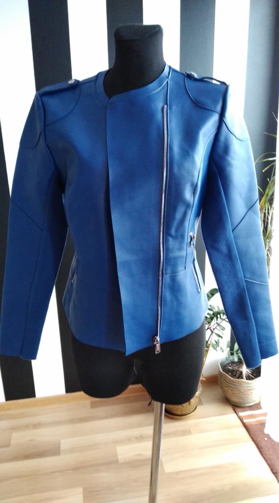 Niebieska, asymetryczna kurtka skórzana z metalowym zamkiem błyskawicznym, prezentowana na czarnym manekinie na drewnianym blacie, z rośliną doniczkową w tle.