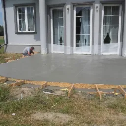 Wylewka betonowa tarasu w trakcie realizacji, widoczne drewniane szalunki i pracownik wygładzający powierzchnię betonu, w tle ściana domu z oknami balkonowymi.