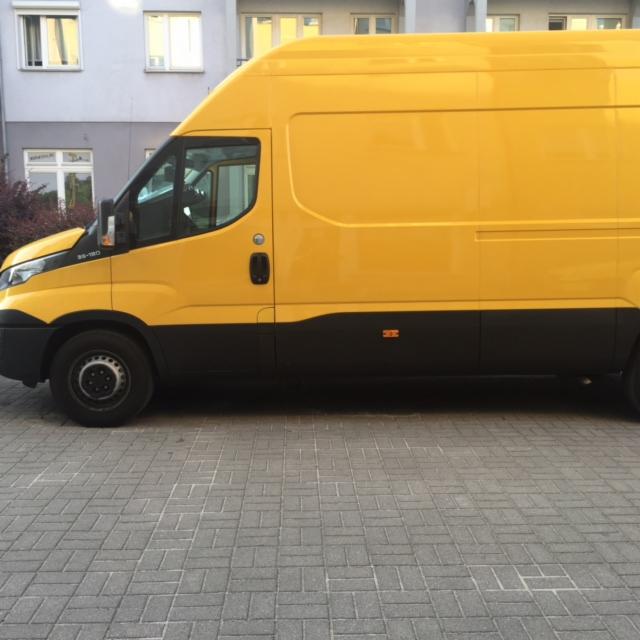 Żółto-czarny bus dostawczy Iveco zaparkowany na brukowanym placu przed budynkiem mieszkalnym.