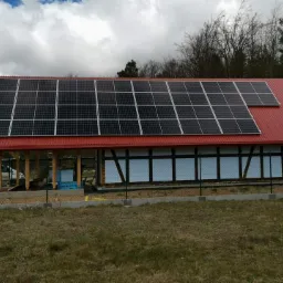 Instalacja paneli słonecznych na czerwonym dachu budynku gospodarczego w trakcie budowy, widoczne materiały izolacyjne i częściowo ukończone ogrodzenie.