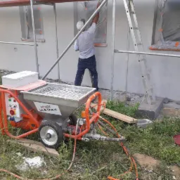 Pracownik w białej koszulce nanosi tynk maszynowy na elewację domu, widoczna maszyna do tynkowania marki Małek M-S MAX, okna zabezpieczone folią, rusztowanie i drabina w tle.