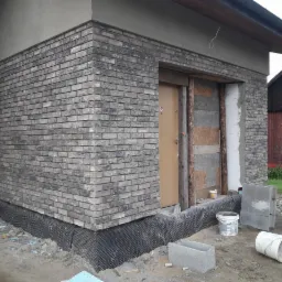 Szara elewacja z cegły klinkierowej w trakcie budowy, widoczne bloczki betonowe, fragment membrany hydroizolacyjnej przy fundamencie, surowe drewno wokół otworu drzwiowego.