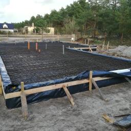 Pro-Builder Paweł Szymański - Przygotowanie zbrojenia fundamentu pod budynek, widoczne drewniane szalunki, czarna folia izolacyjna, siatka z prętów zbrojeniowych oraz wystające rury instalacyjne. W tle zabudowa jednorodzinna...