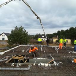 Pro-Builder Paweł Szymański - Prace na budowie w Lipnie: Ekipa budowlana w kamizelkach odblaskowych przygotowuje zbrojenie stropu z widocznymi instalacjami, z betonową pompą w tle i domami w oddali.