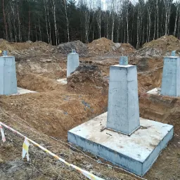 Betonowe słupy fundamentowe na placu budowy, widoczne wykopane rowy i taśmy ostrzegawcze, w tle las.